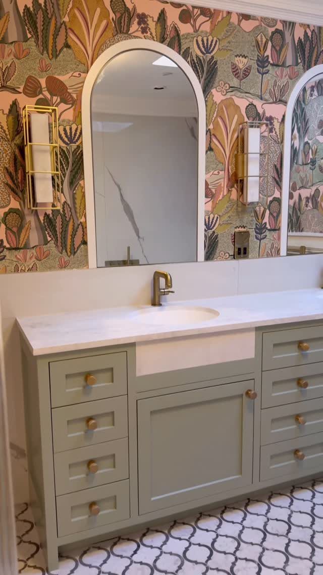 A light green bathroom vanity with marble countertops and arched mirrors against patterned wallpaper.