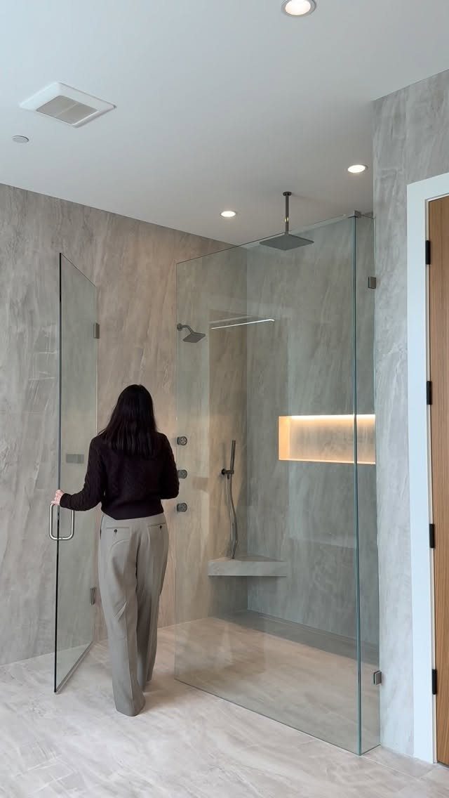 A woman standing in front of a modern frameless glass walk-in shower with grey tiles.