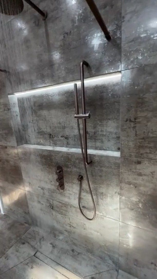 Grey stone tiled shower wall with a lit recessed niche and dark metal handheld shower fixture.