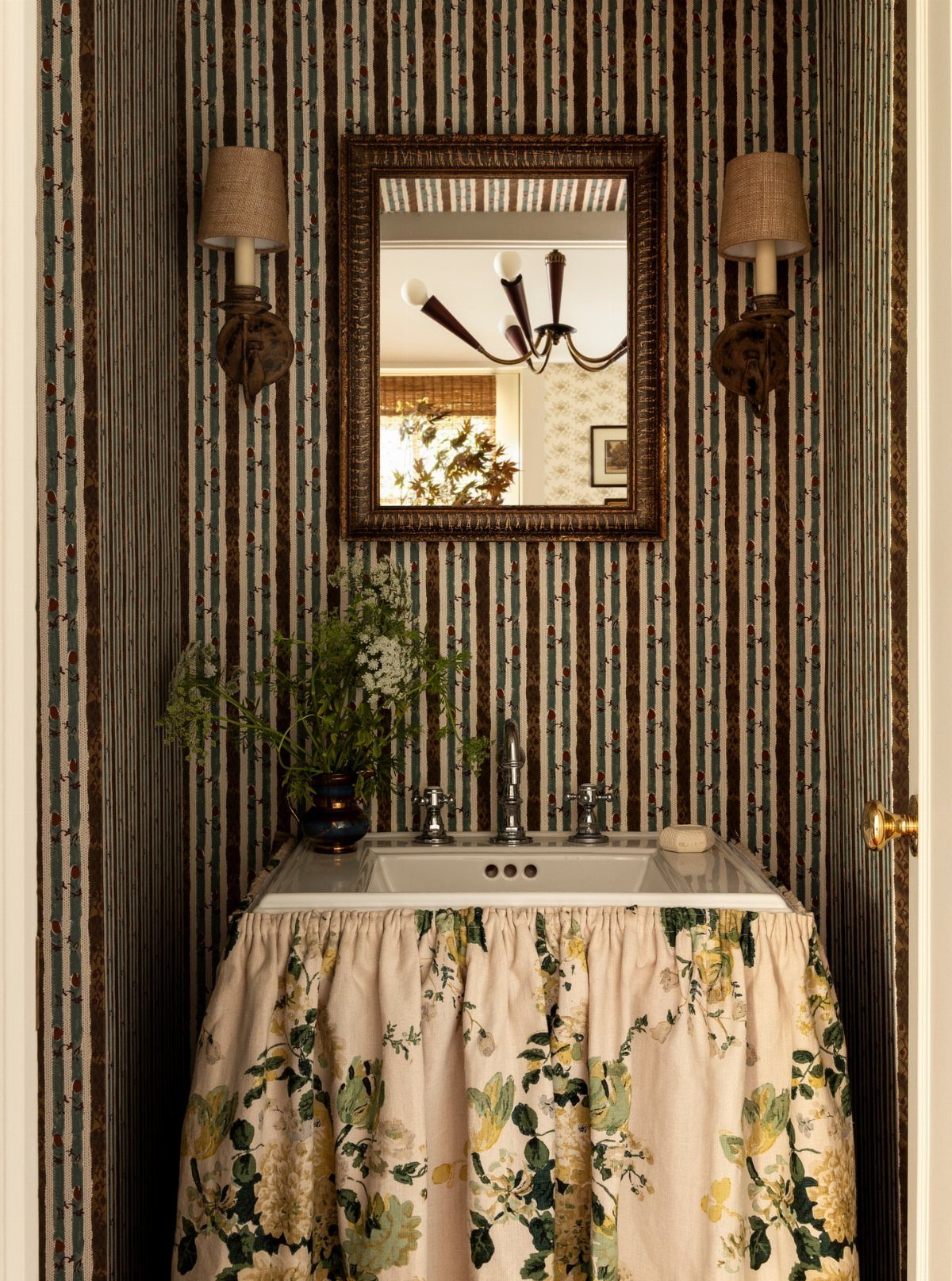 A white bathroom sink featuring a decorative floral fabric skirt against striped wallpaper.