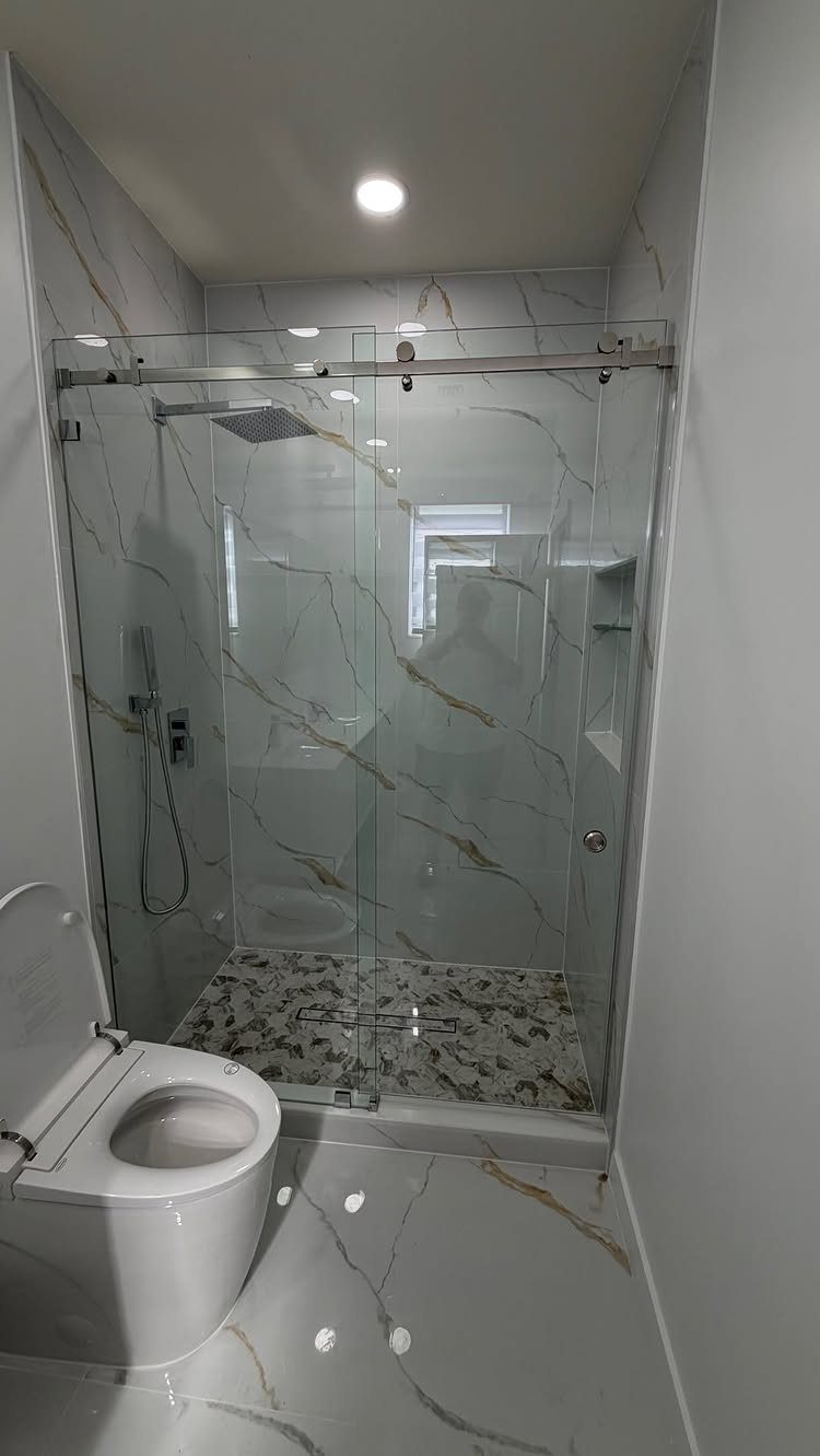 A narrow bathroom with white marble tiles and a glass sliding shower partition.