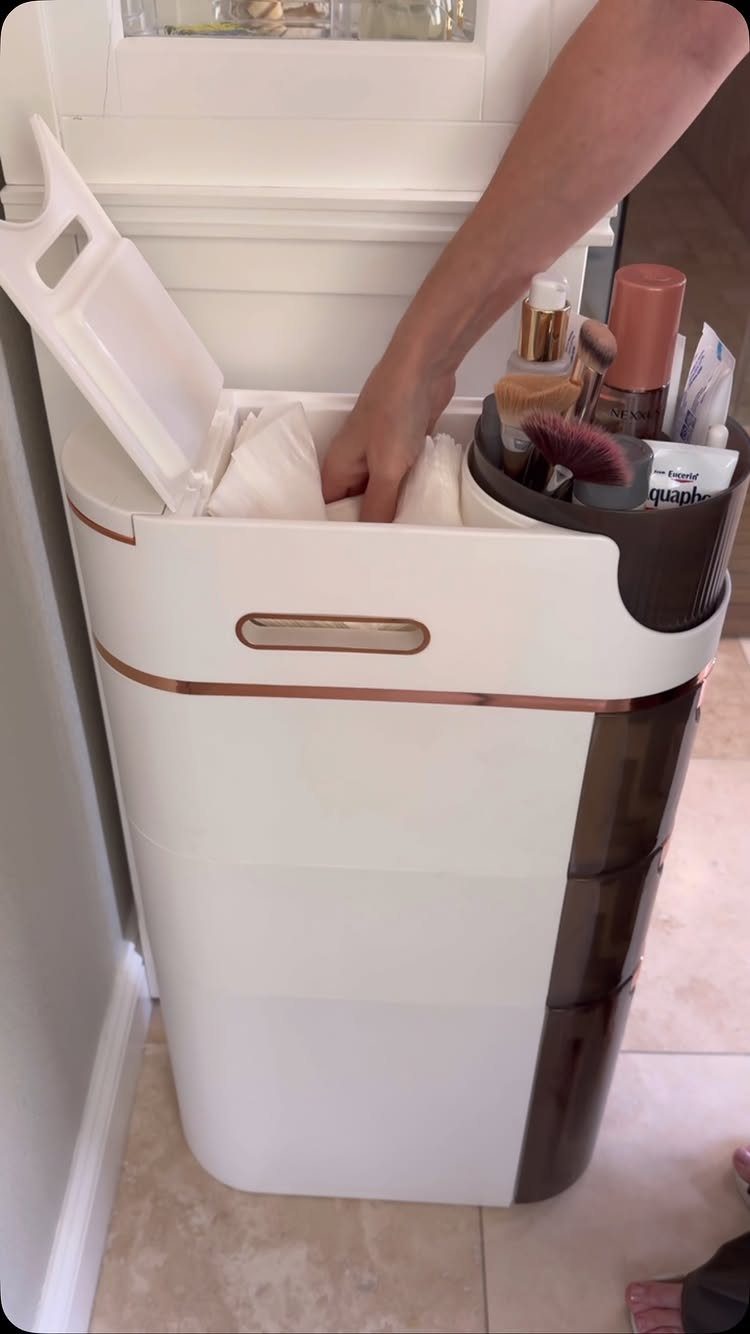 A person organizing toiletries inside a slim, white vertical storage cart on a tiled floor.