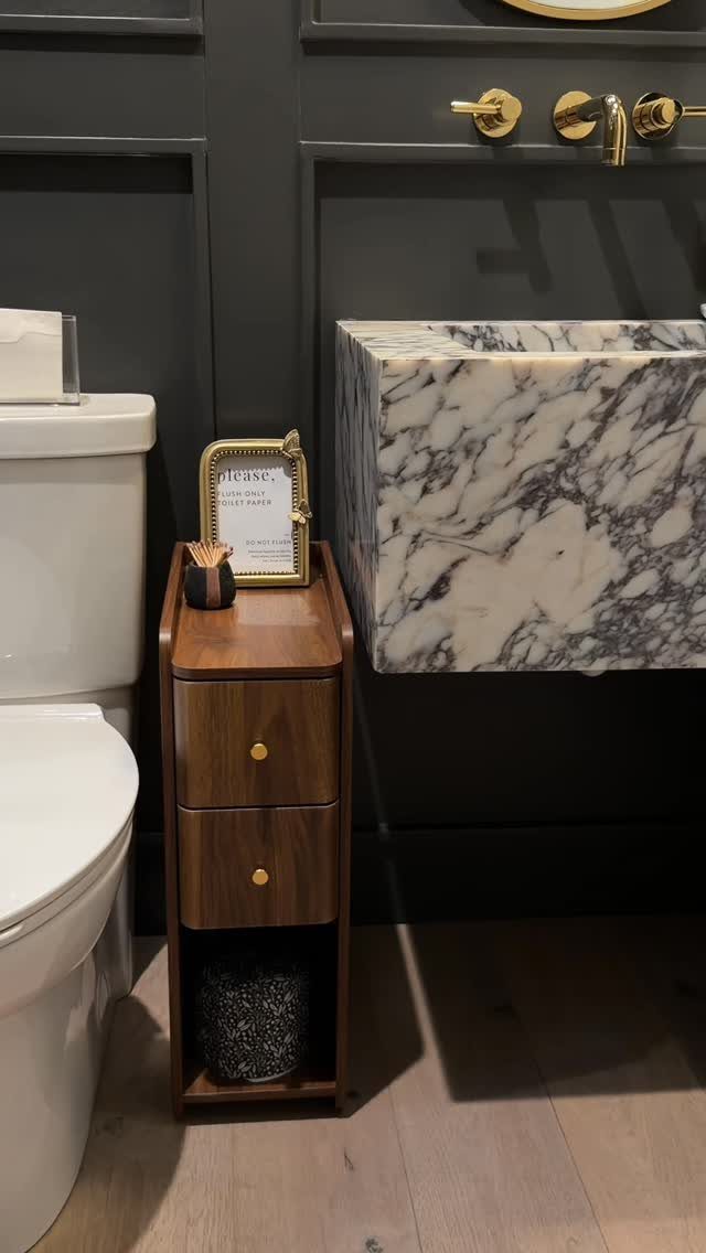 A narrow wooden two-drawer storage unit stands between a white toilet and a marble sink.