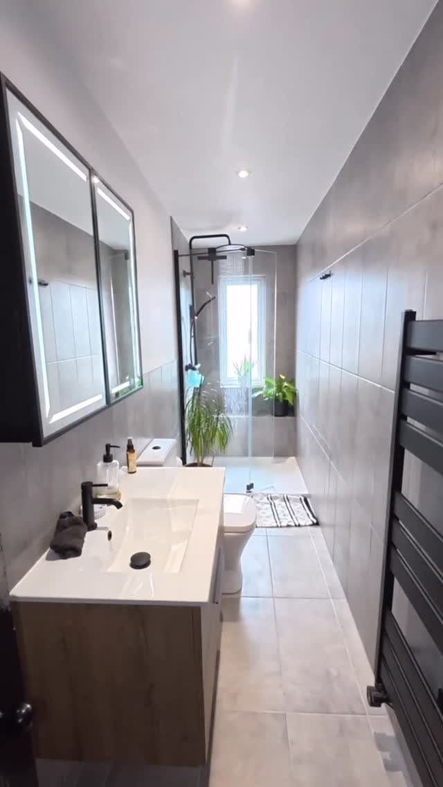 A narrow modern bathroom with grey tiles, a wooden vanity, and a glass shower enclosure.