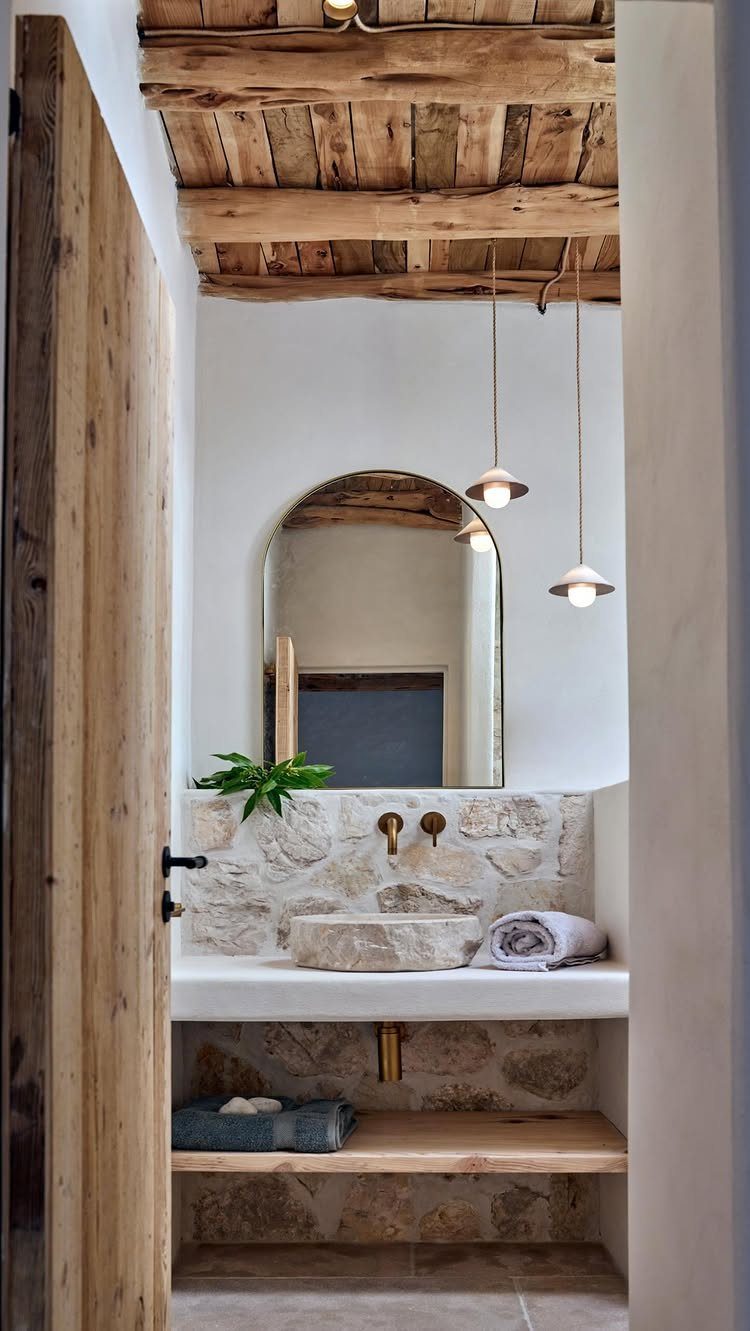 A rustic bathroom with stone walls, an arched mirror, and a wooden beam ceiling.