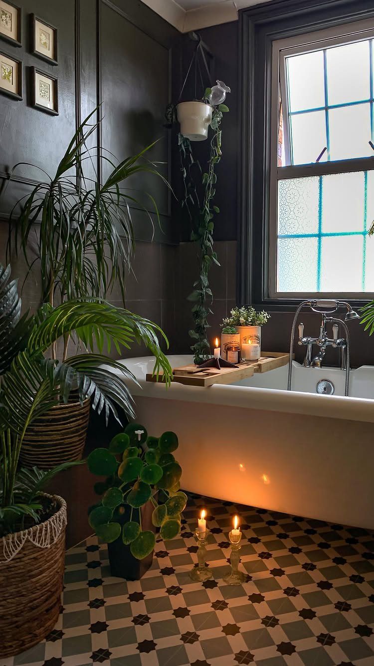 A white bathtub surrounded by lush green plants and glowing candles in a dark bathroom.