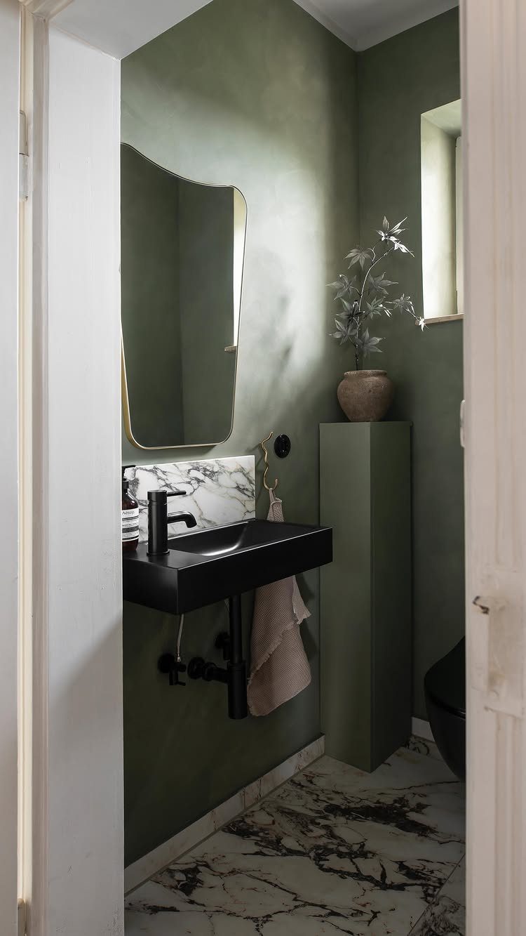A uniquely shaped gold-framed mirror hanging above a black sink in a green bathroom.