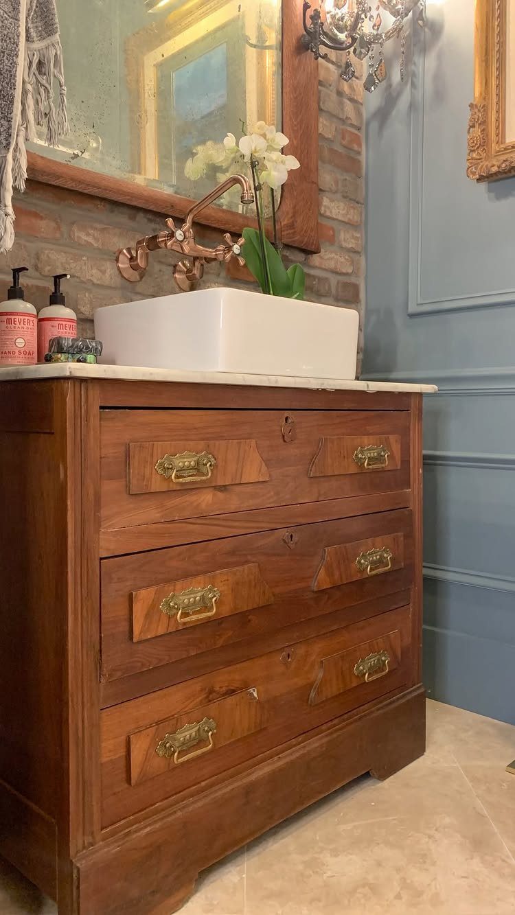 Antique wooden dresser vanity with a white vessel sink and copper faucet against a brick wall.