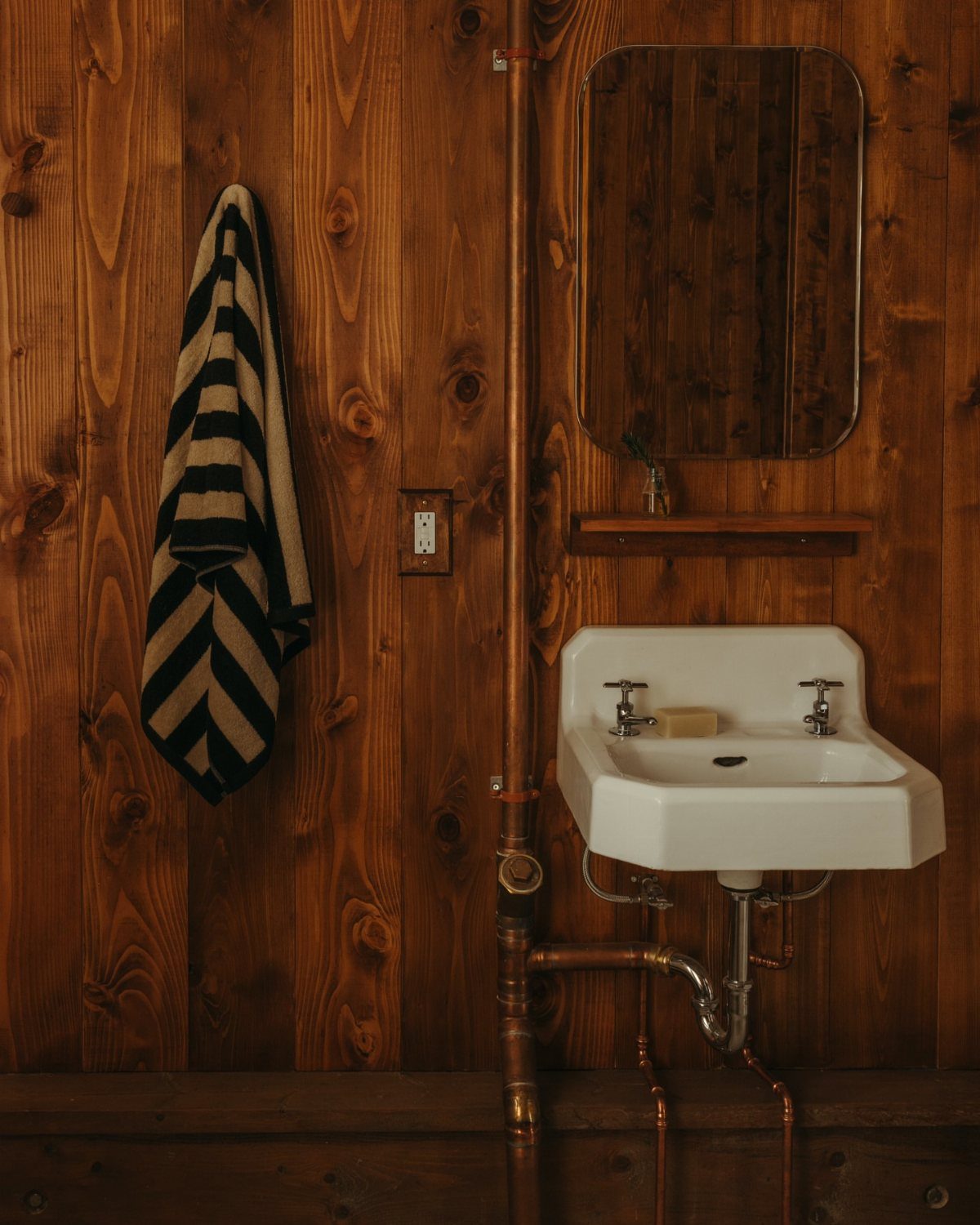 White antique wall-mounted sink with copper pipes against a vertical wood paneled wall.