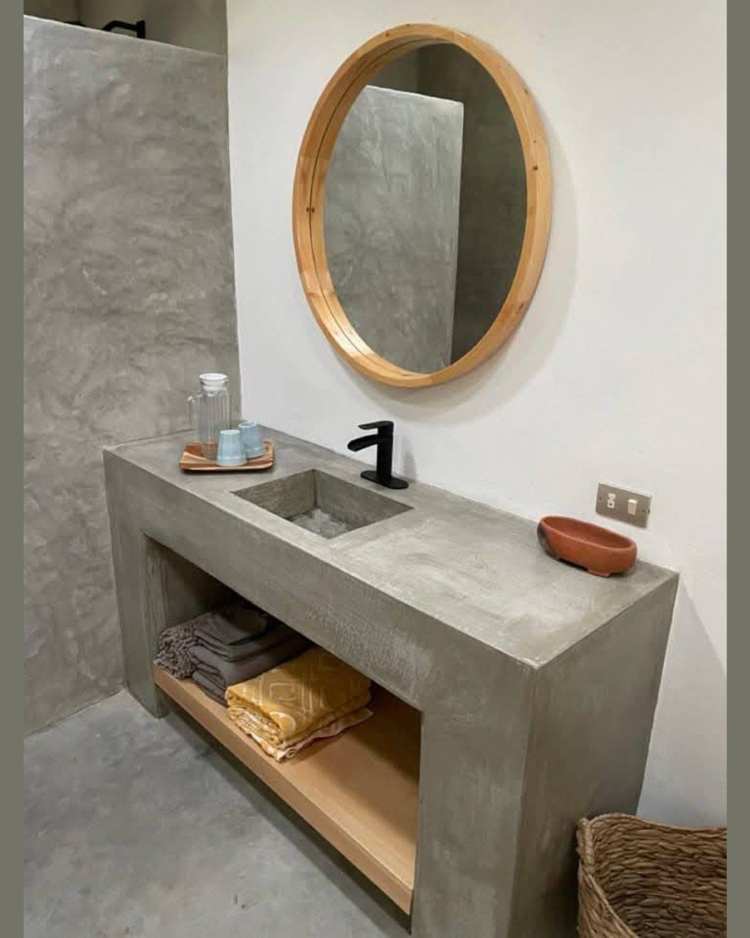 A grey cement vanity with a built-in sink, black faucet, and round wooden mirror.