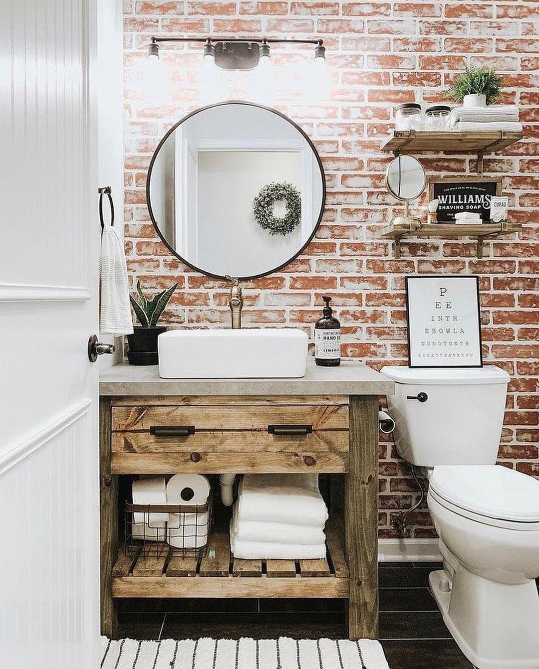 A rustic bathroom with a red brick wall, wooden vanity, white vessel sink, and round mirror.
