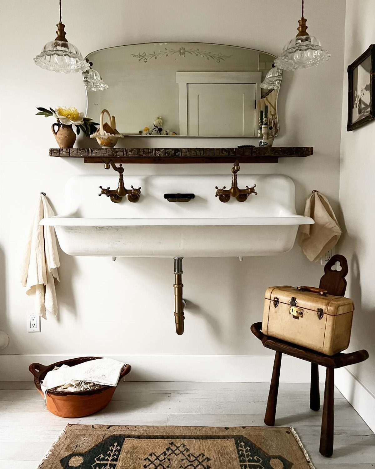 A white wall-mounted trough sink with brass faucets and a wooden shelf above it.