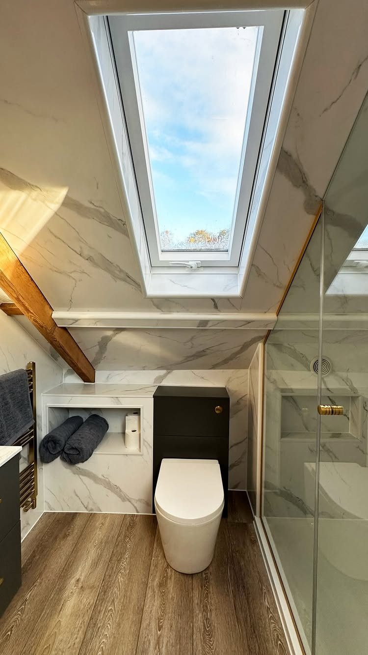 A modern bathroom with marble walls, wood flooring, and a large skylight in a vaulted ceiling.