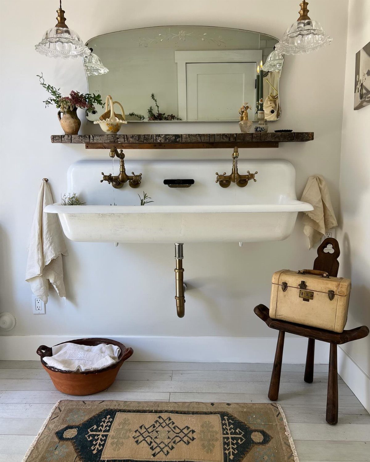 A white wall-mounted trough sink with brass fixtures and vintage decor in a bright bathroom.