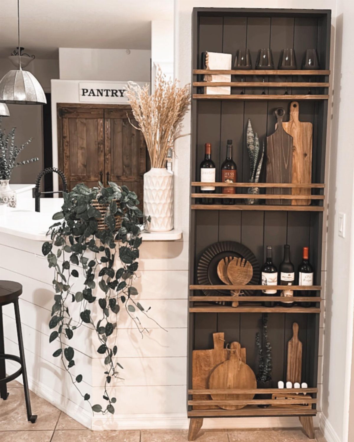 A tall dark grey wooden shelf unit holding wine bottles, glassware, and various wooden cutting boards.