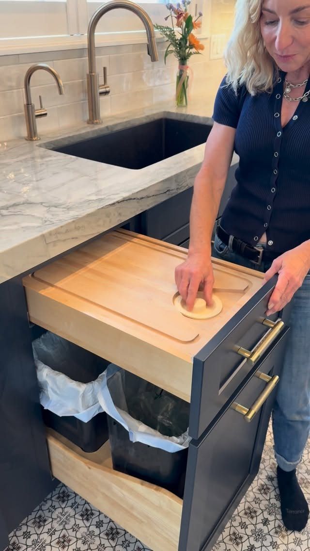 A woman pulling out a kitchen cabinet drawer containing a wooden cutting board and trash bins.