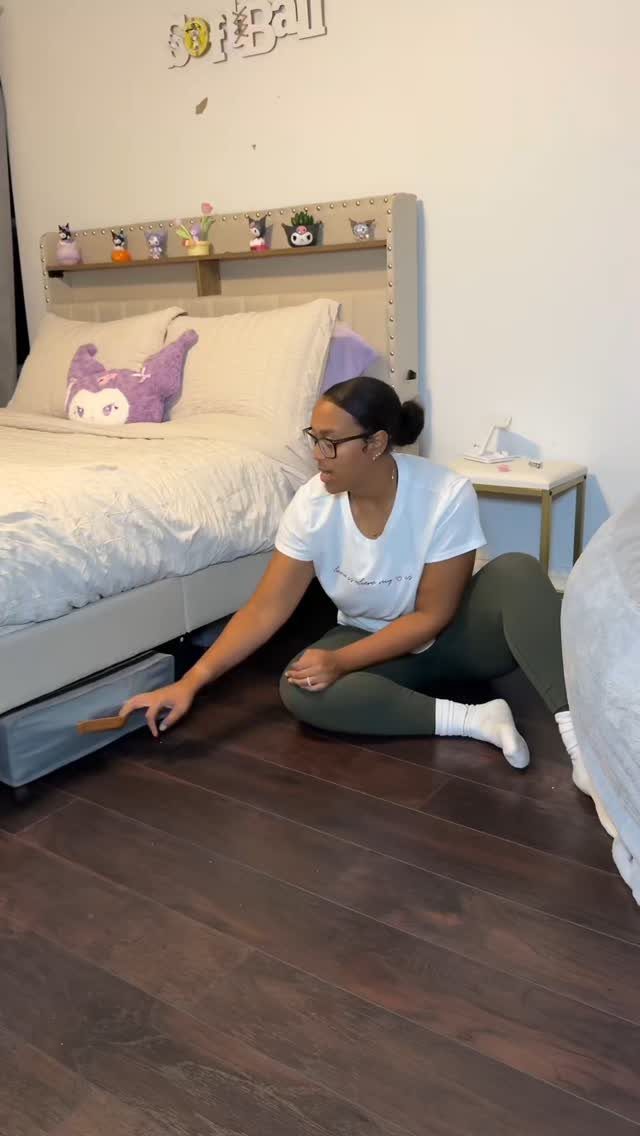 A woman reaches for a large storage box placed underneath a bed on a dark floor.