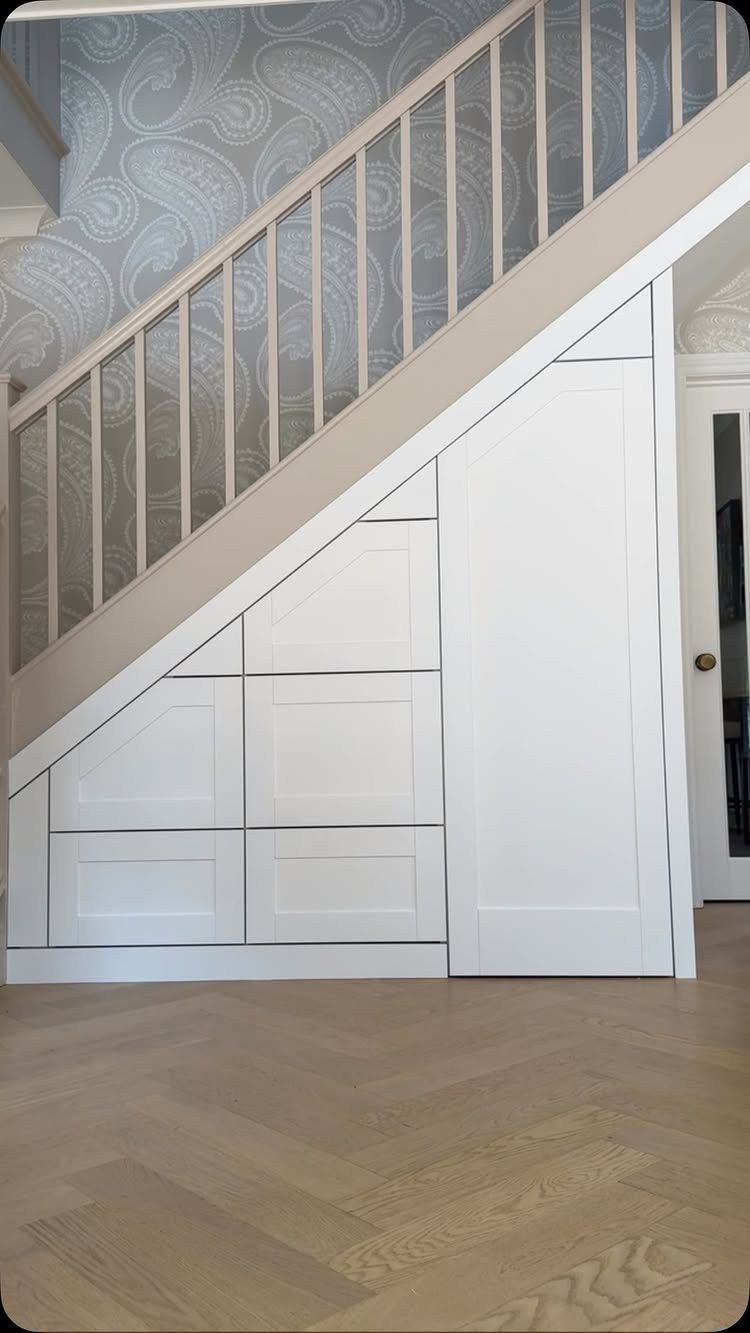 White built-in storage drawers and cabinets installed underneath a wooden staircase with paisley wallpaper.