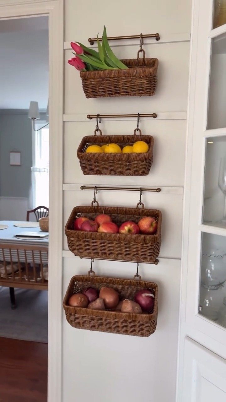 Four wicker baskets hanging on a white wall containing tulips, lemons, apples, and onions.
