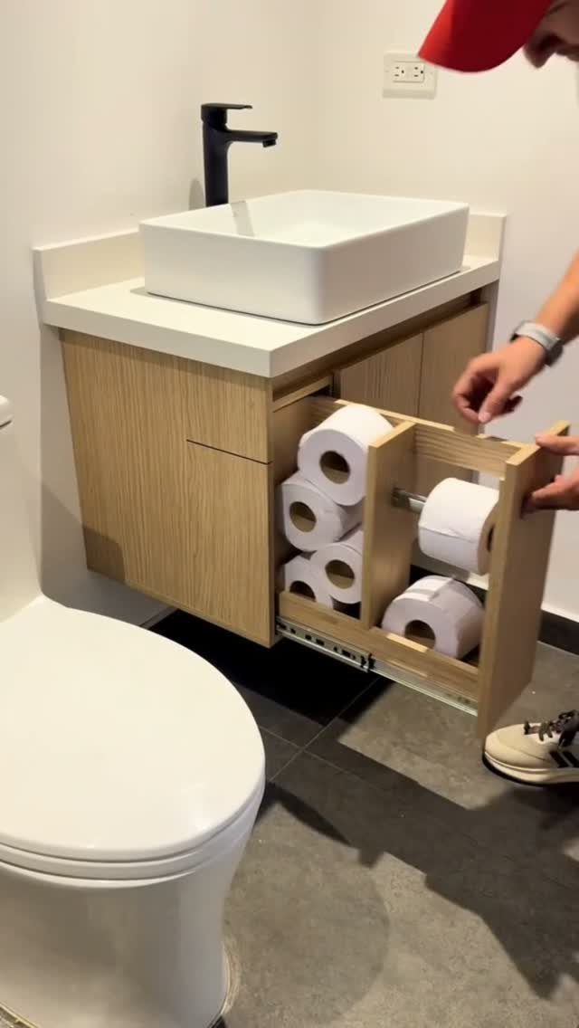 A person pulls out a wooden drawer from a bathroom cabinet filled with toilet paper rolls.