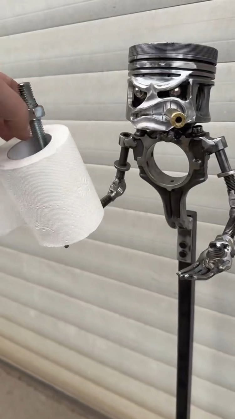A skull-shaped piston engine part sculpture holding a roll of white toilet paper.