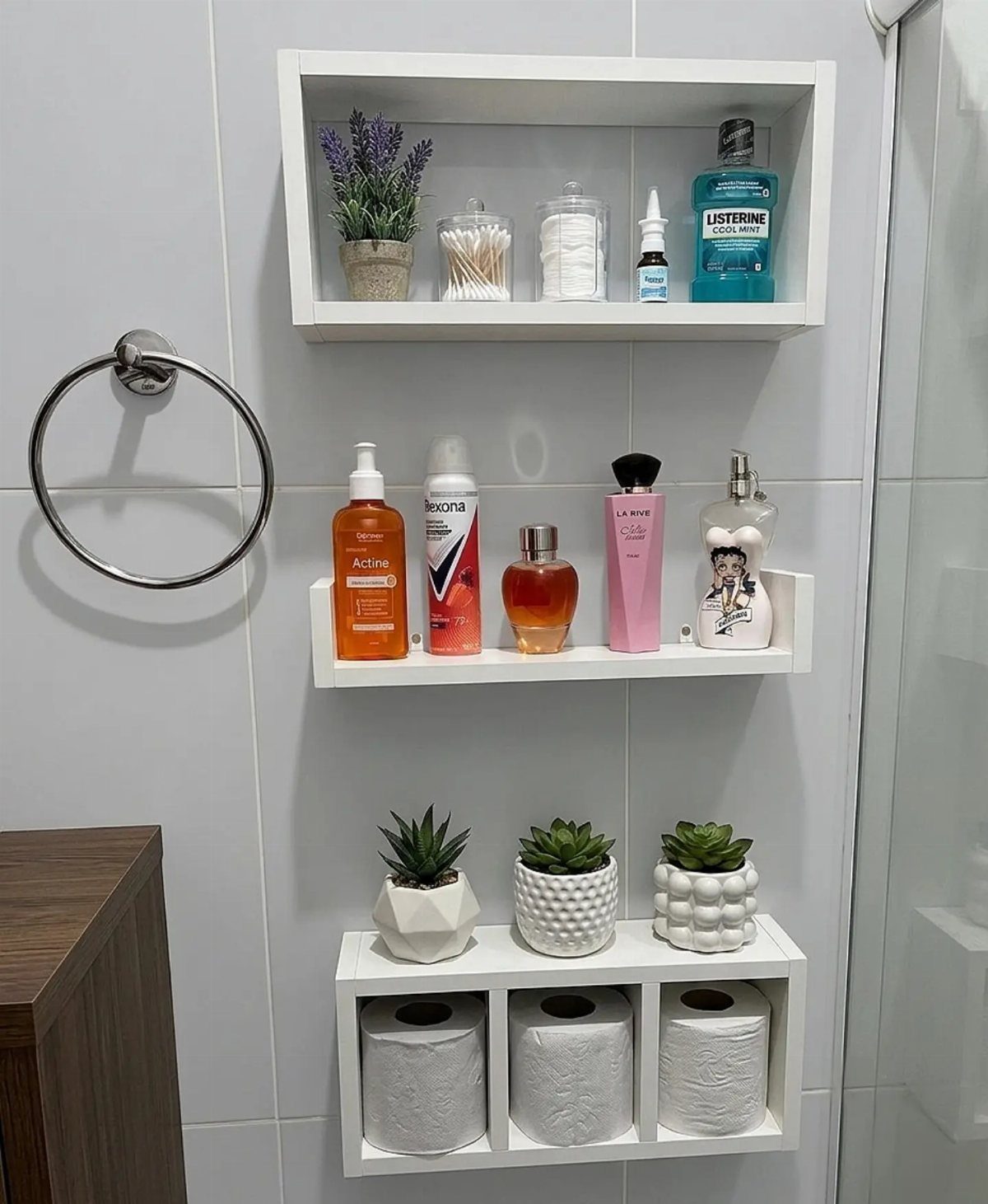 Three white floating shelves mounted on a tiled wall holding toiletries, perfumes, and toilet paper rolls.