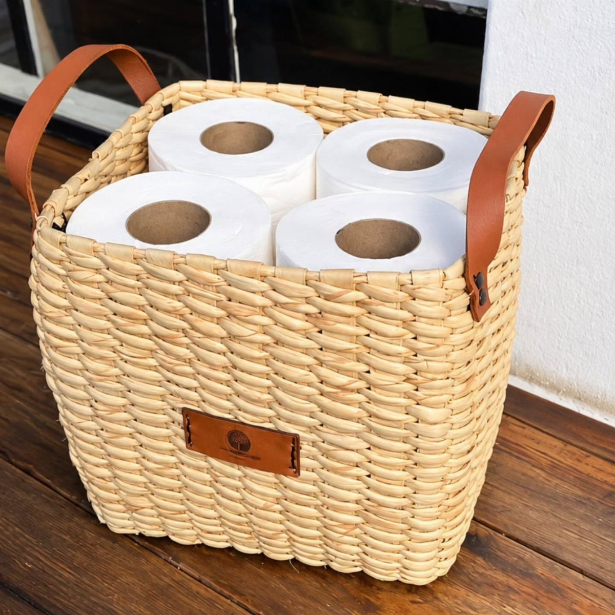 A woven seagrass basket with leather handles containing four rolls of white toilet paper.