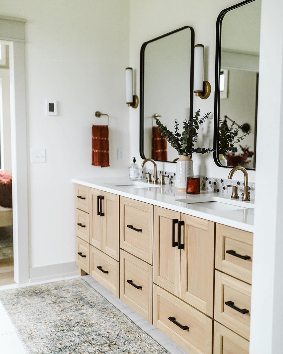 Light wood bathroom vanity with black cabinet pulls and gold faucets and fixtures.