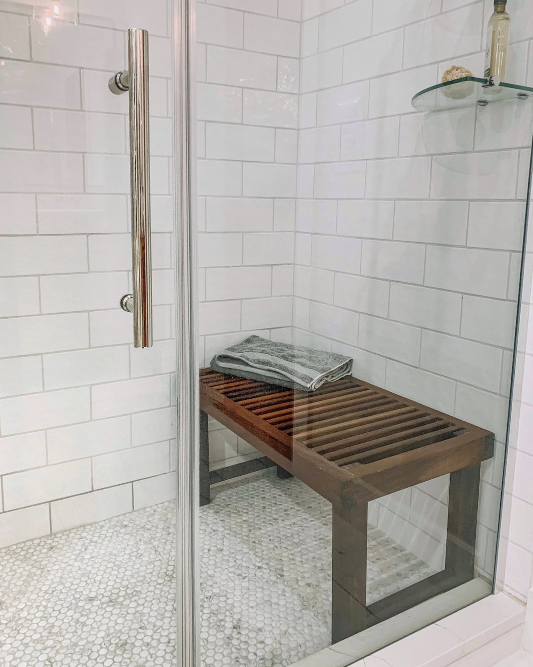 A wooden slatted bench placed inside a white tiled shower with pebble flooring.