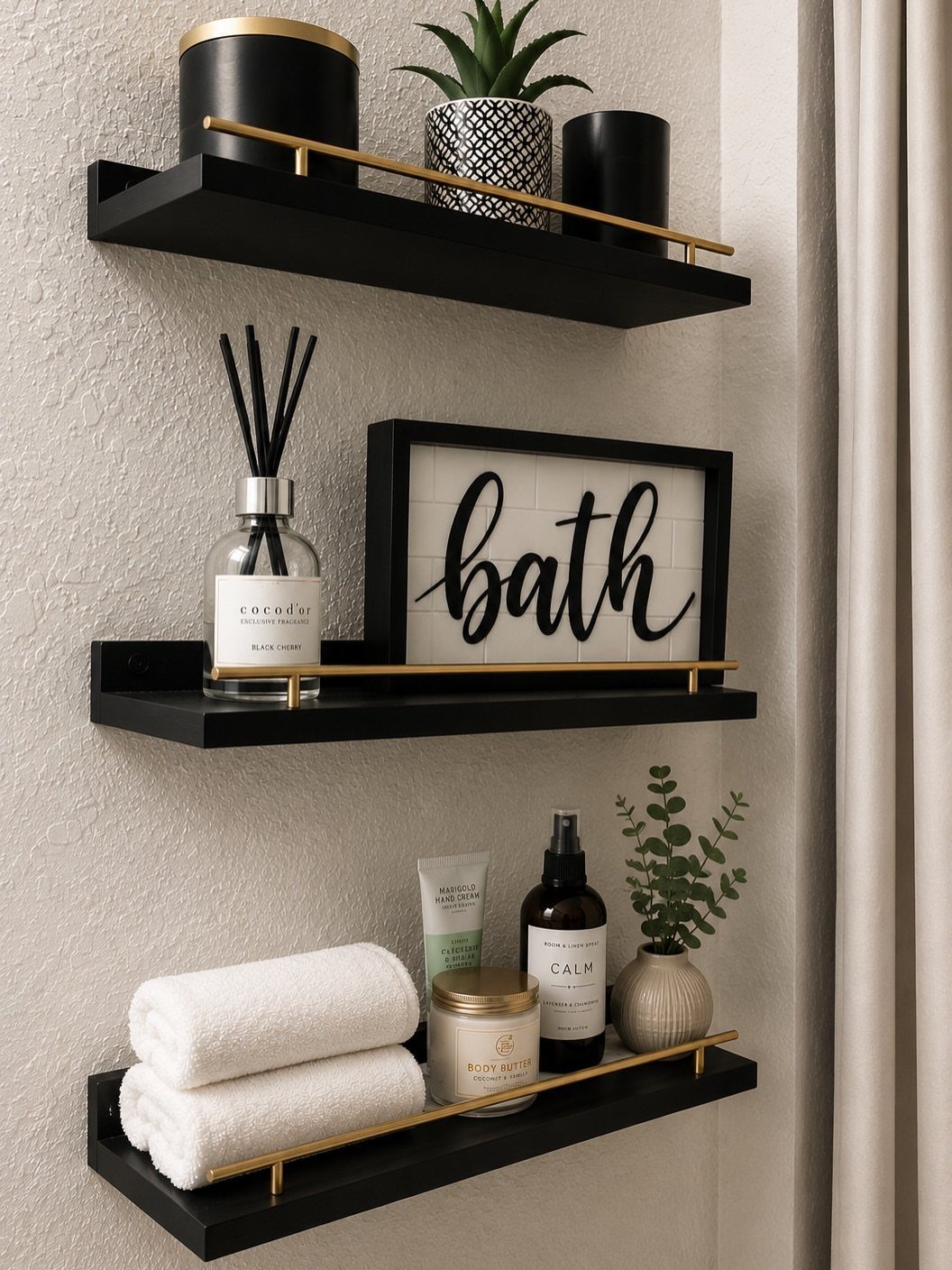 Three black floating shelves with gold accents holding bathroom decor and towels on a wall.