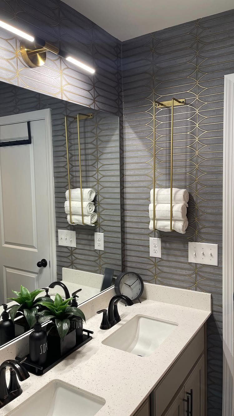 Bathroom vanity with white countertop, black faucets, and patterned grey wallpaper with gold accents.