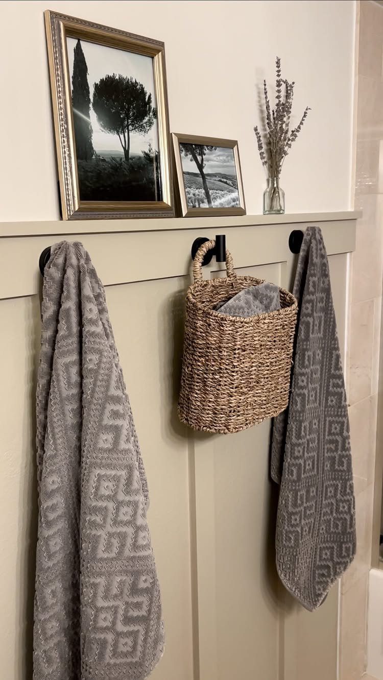 Grey patterned towels and a woven basket hanging on hooks against a beige wainscoted wall.