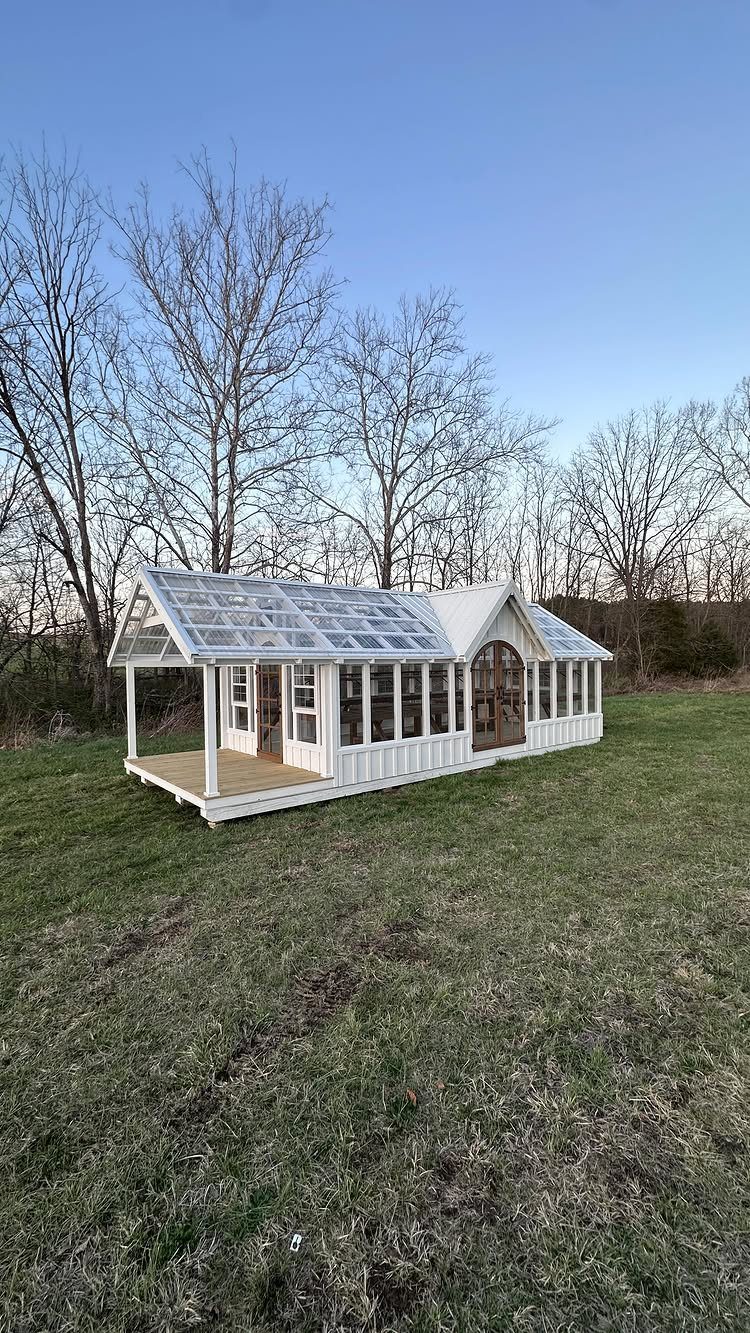 A white wooden glass garden shed with an integrated wooden porch on a grassy lawn.
