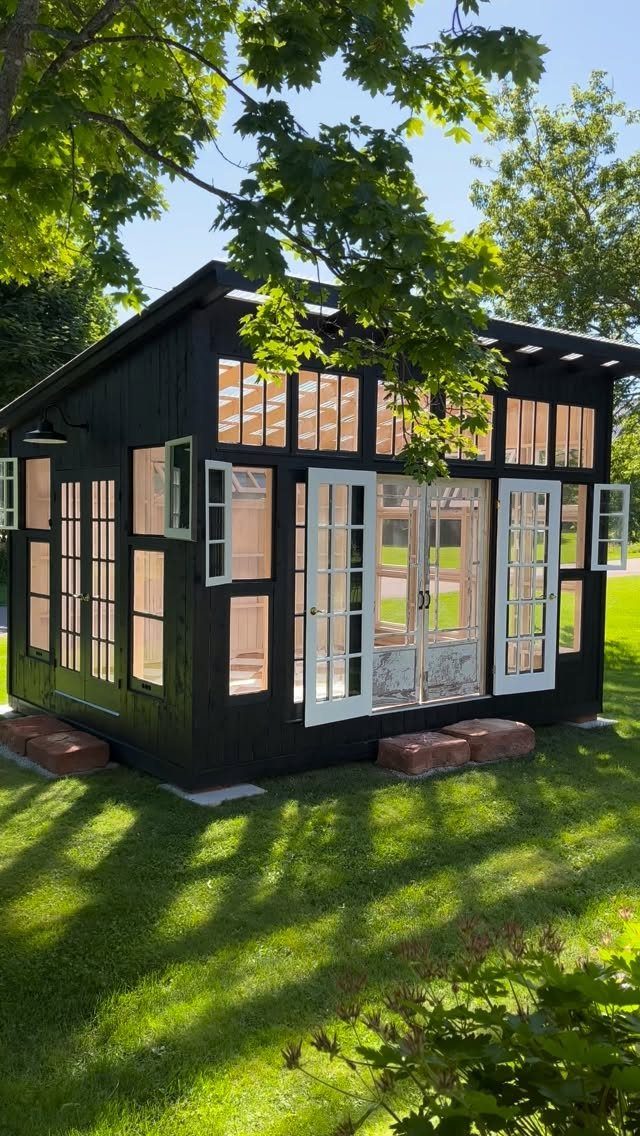 A black wooden garden shed with many large glass windows and white doors on a lawn.
