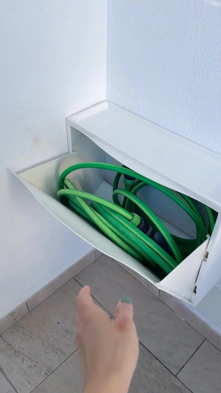 A white wall-mounted storage box holding a coiled green garden hose against a white wall.