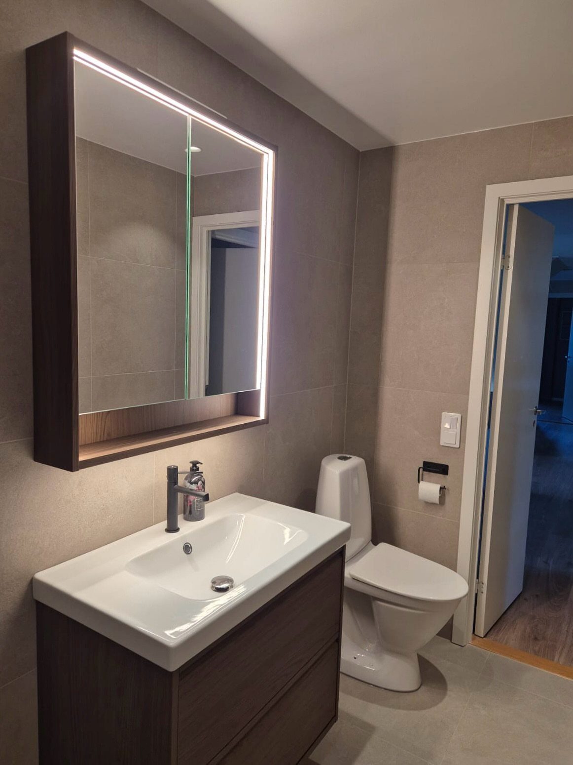 A dark wood bathroom vanity with a white sink sits below a large illuminated mirror.