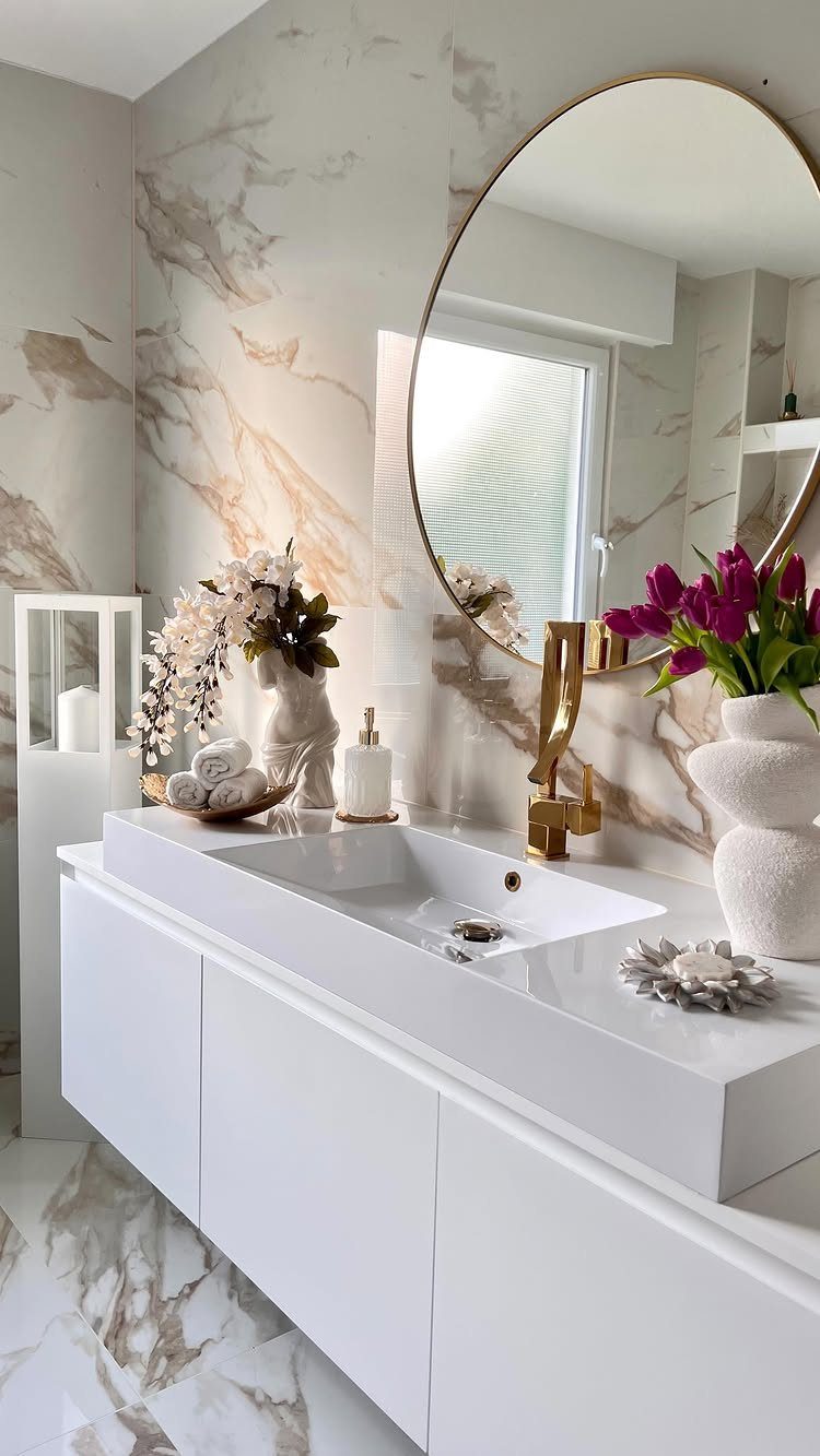 White marble bathroom vanity with a gold faucet, round mirror, and floral decorations.