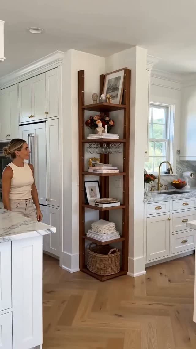 Tall wooden corner shelving unit filled with decor and baskets in a bright white kitchen.