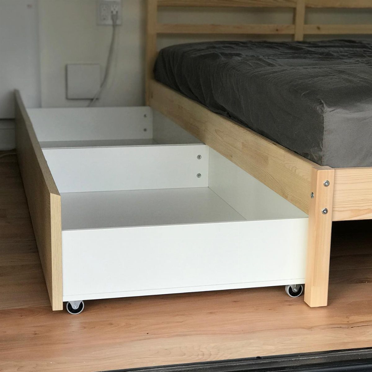 A white rolling storage drawer being pulled out from under a light wood bed frame.