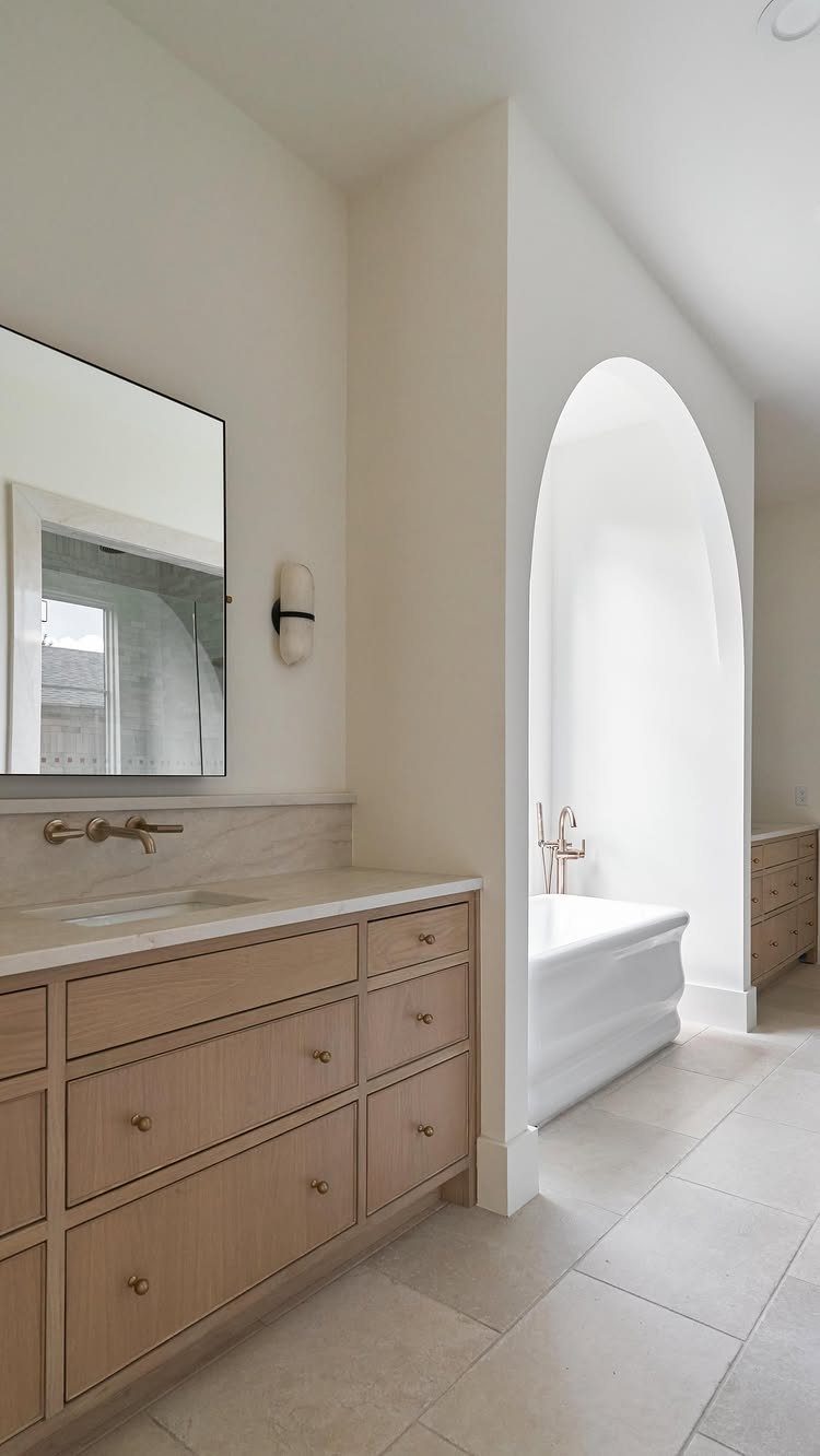 A light wood bathroom vanity with a large mirror next to an arched alcove containing a bathtub.