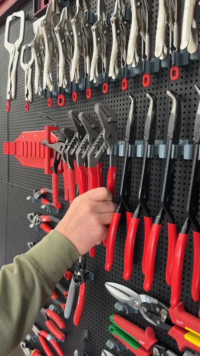 A hand reaches to grab red-handled pliers from a black pegboard filled with various tools.