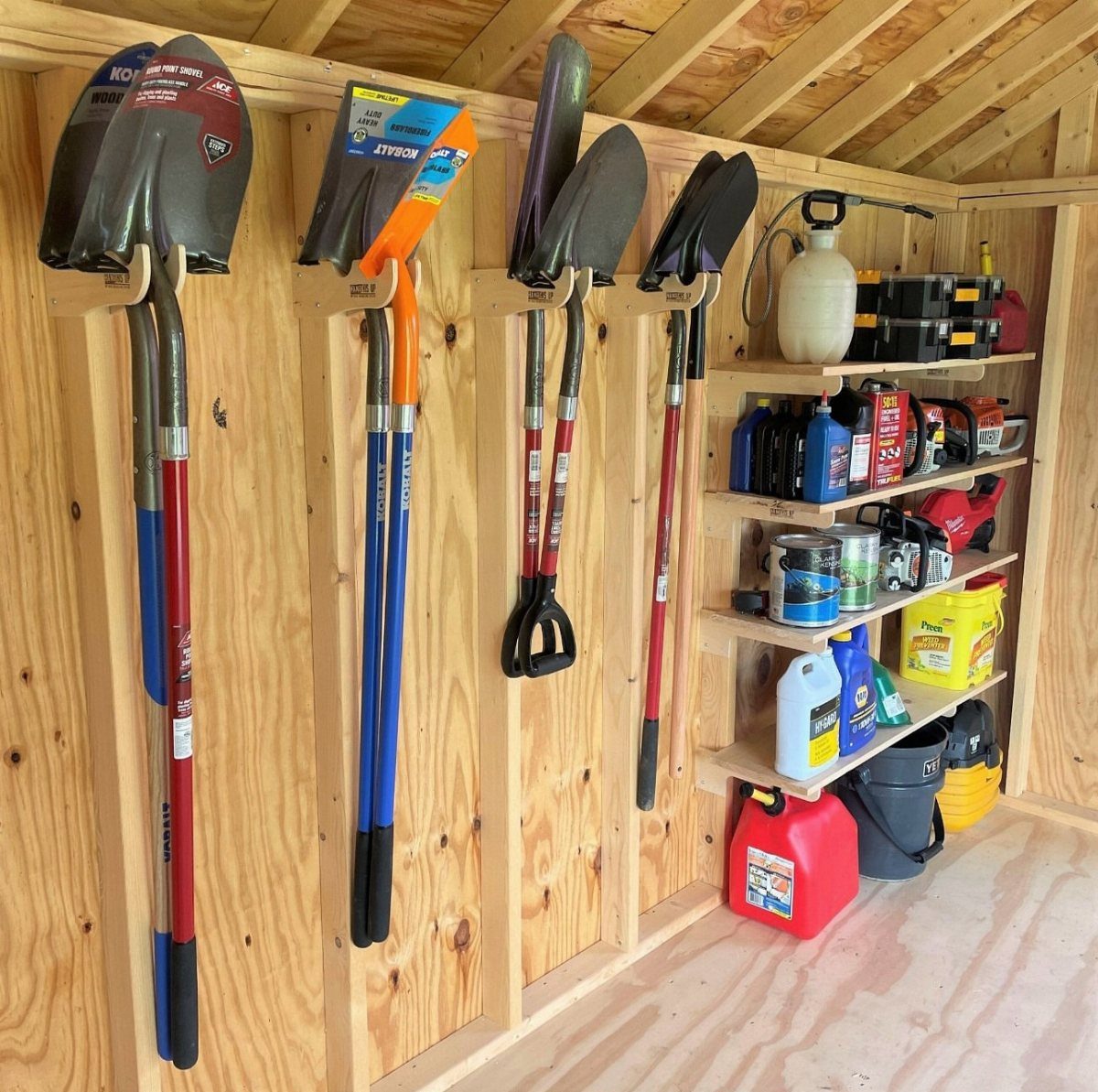 Garden tools hanging on wooden wall studs next to shelves filled with various containers.