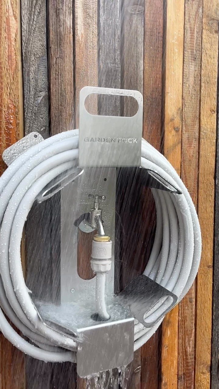 A white garden hose coiled on a metal wall-mounted reel against a wooden fence.
