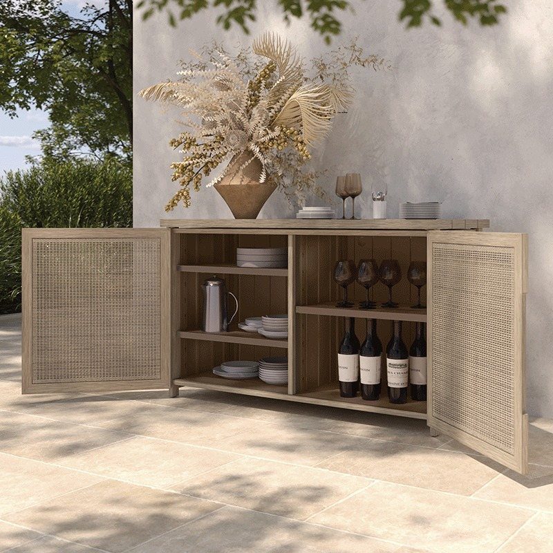 A light wood outdoor bar credenza with cane doors holding wine, glassware, and plates.