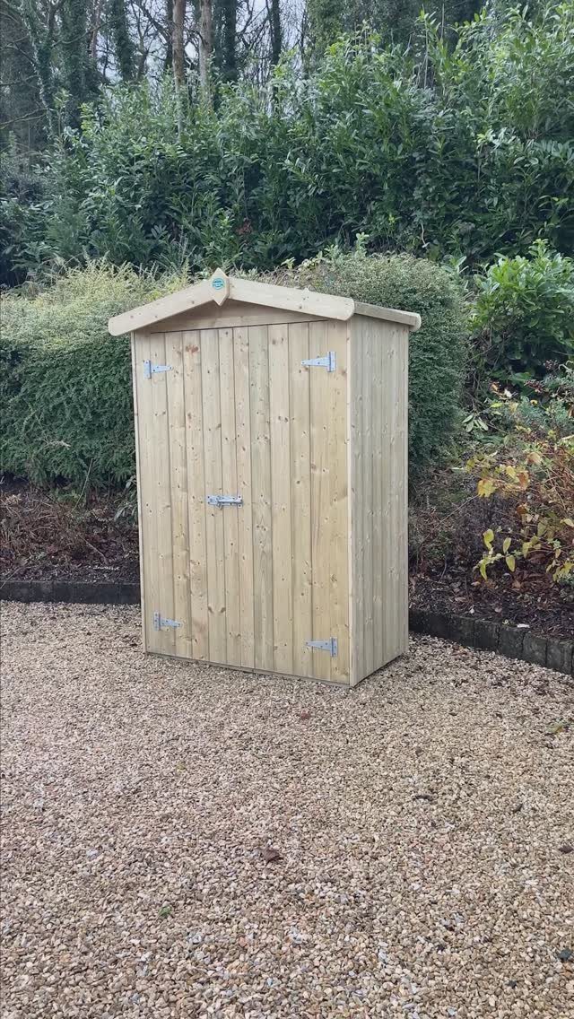 A small wooden garden shed sits on a gravel path surrounded by green bushes.