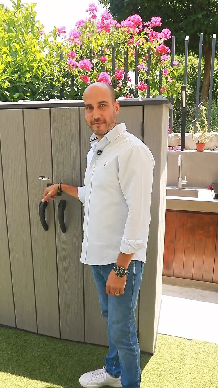 A man standing beside a large grey vertical outdoor storage cabinet on a sunny patio.