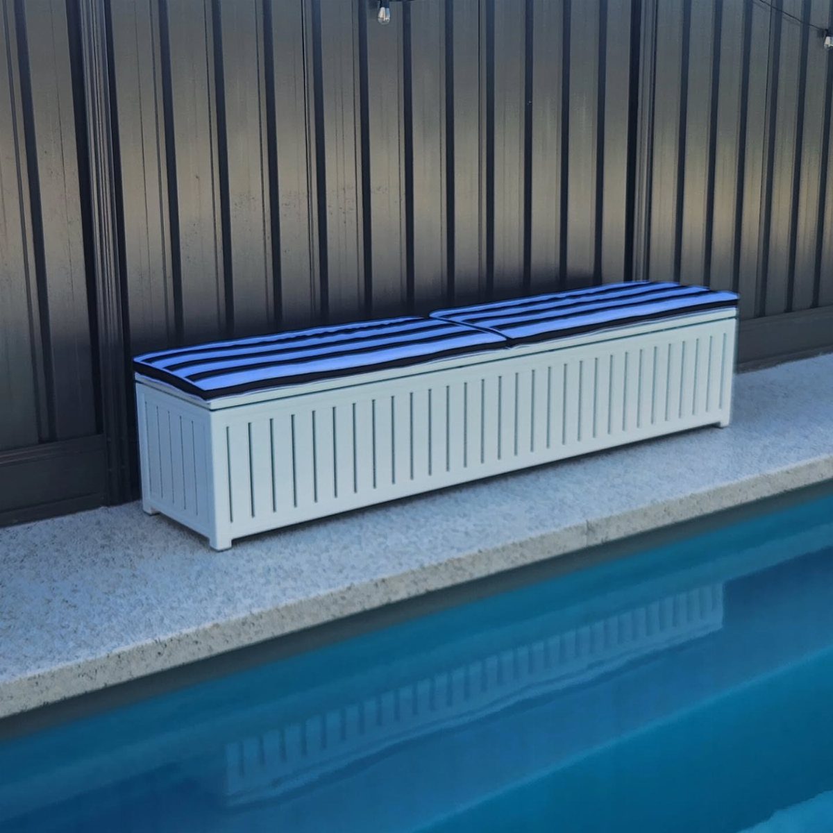 A white slatted deck box with a blue striped lid sits beside a swimming pool.