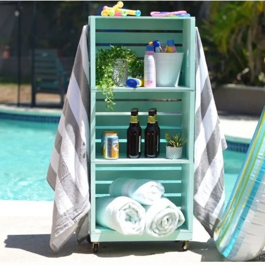 A light blue wooden rolling cart with towels and drinks sits next to a swimming pool.