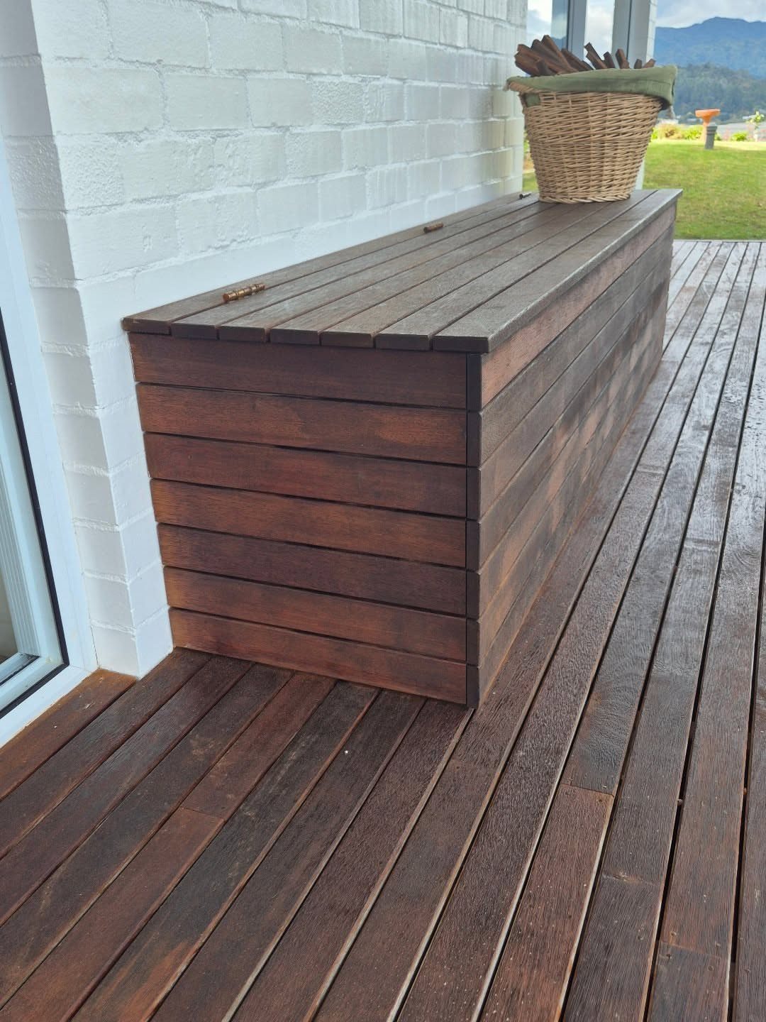 A long dark wood storage bench sits on a wooden deck against a white brick wall.