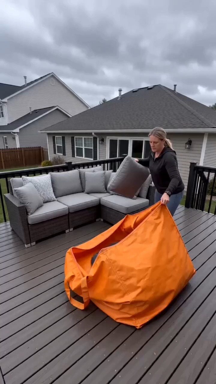 A woman placing grey outdoor cushions into a large orange waterproof storage bag on a wooden deck.